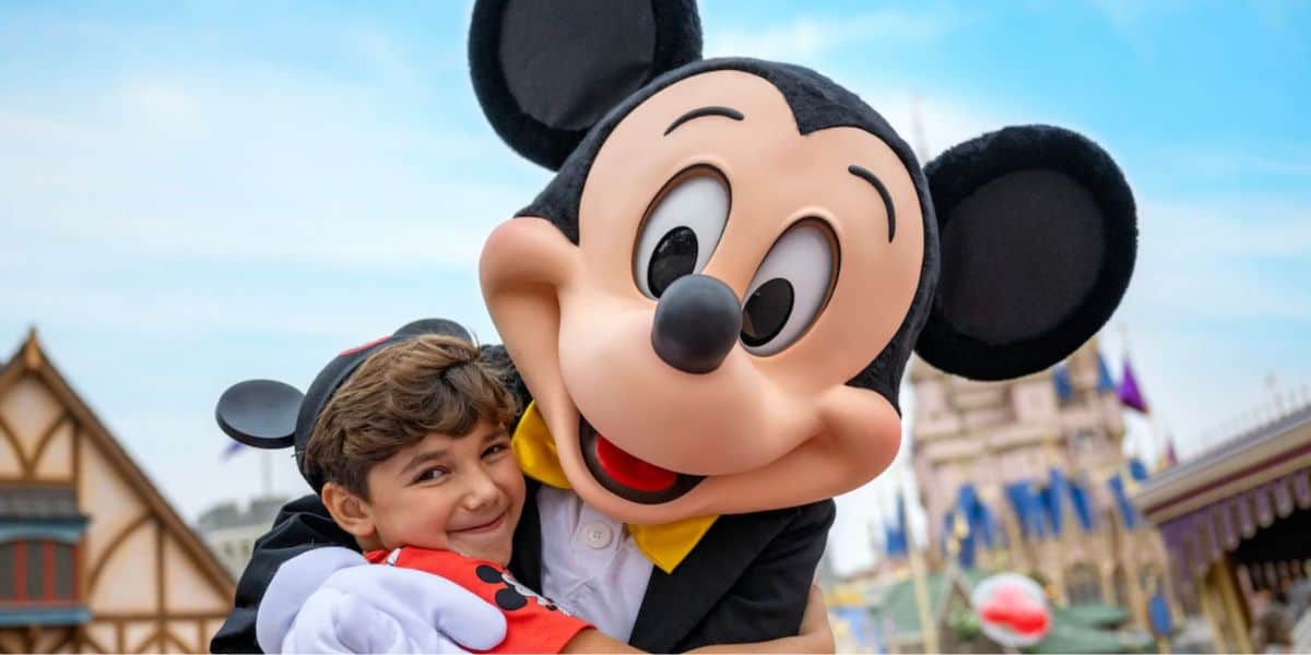 El pequeño bebé de Mickey sonríe mientras abraza la ropa del parque Mickey Mouse, con sus coloridos edificios y banderas.