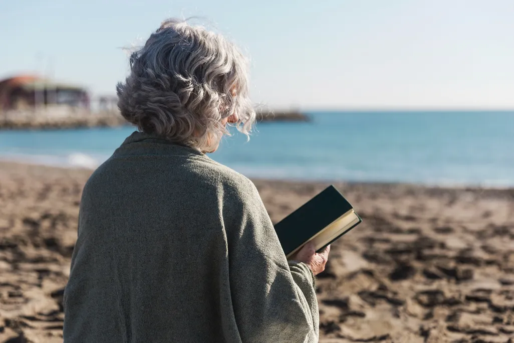 Una mujer de pelo blanco con un libro en la playa.