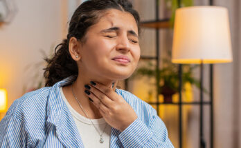 A woman holding her throat in pain while sitting at her desk at home.