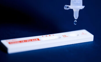 A COVID-19 test lying flat with a droplet falling from a syringe.