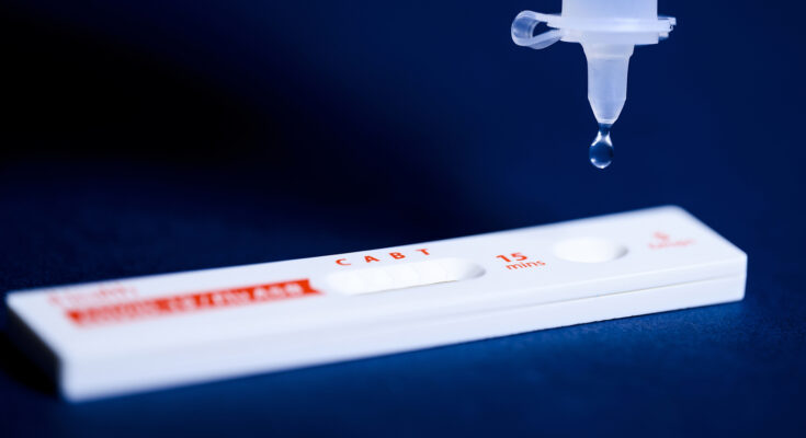 A COVID-19 test lying flat with a droplet falling from a syringe.