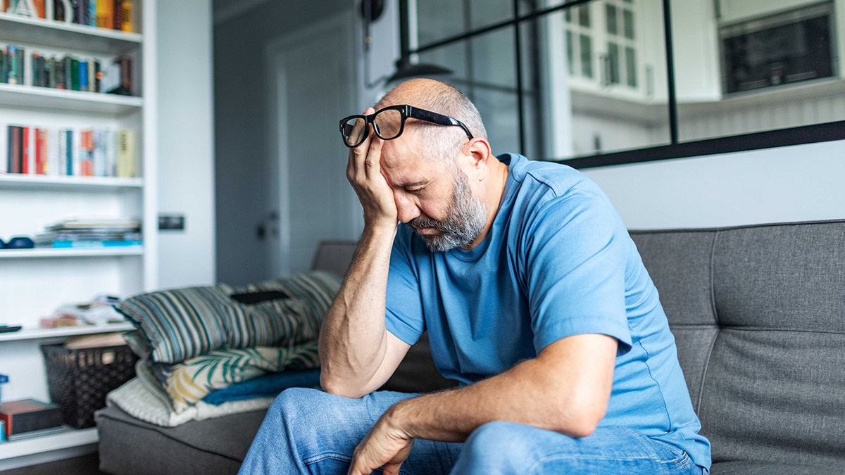 Hombre de mediana edad sentado en un sofá con la cabeza gacha, luciendo estresado o cansado en casa.