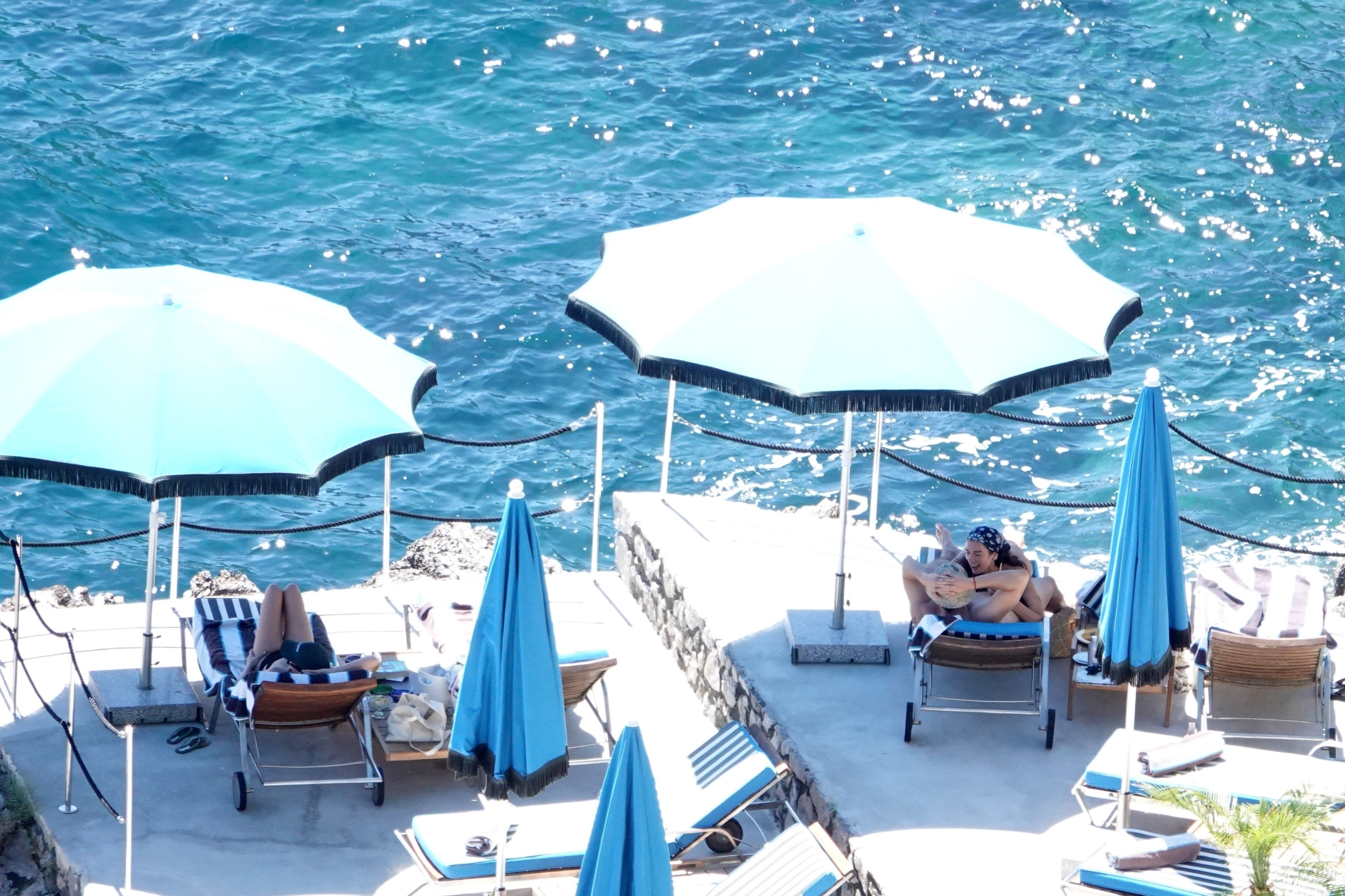 Vista aérea de dos personas abrazadas en una tumbona en una terraza de piedra rodeada de sombrillas azules, con el mar turquesa detrás.